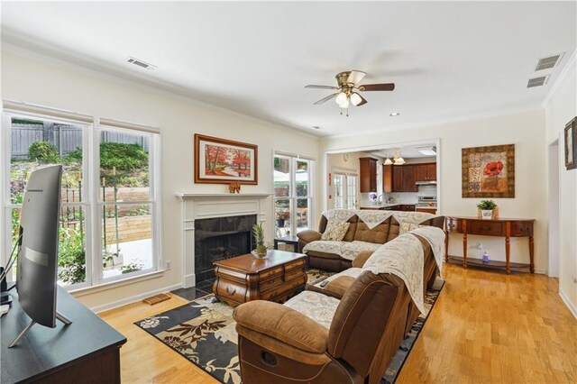 living area with ceiling fan, visible vents, light wood finished floors, a tiled fireplace, and crown molding