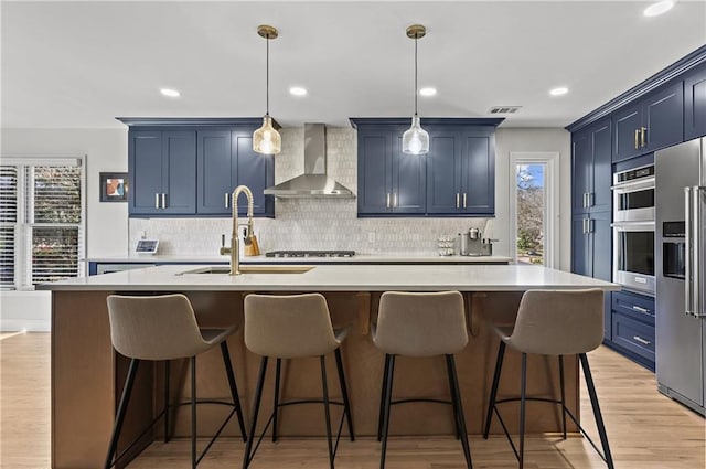 kitchen with blue cabinetry, wall chimney range hood, a wealth of natural light, stainless steel appliances, and a sink