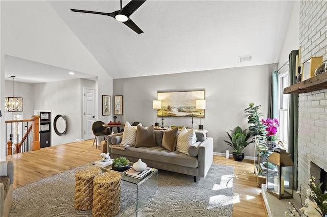 living area featuring light wood-type flooring, visible vents, high vaulted ceiling, ceiling fan with notable chandelier, and a brick fireplace