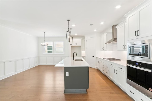 kitchen featuring white cabinets, hanging light fixtures, an island with sink, wall chimney exhaust hood, and stainless steel appliances