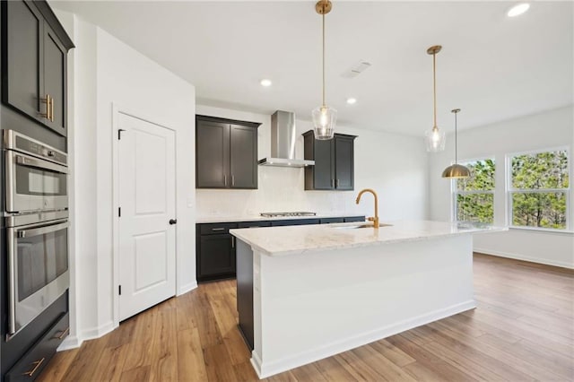 kitchen with an island with sink, wall chimney range hood, pendant lighting, and a sink