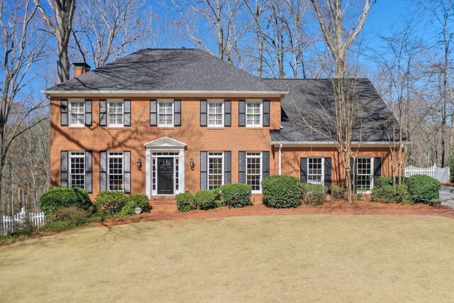 colonial inspired home with brick siding, roof with shingles, a chimney, and fence