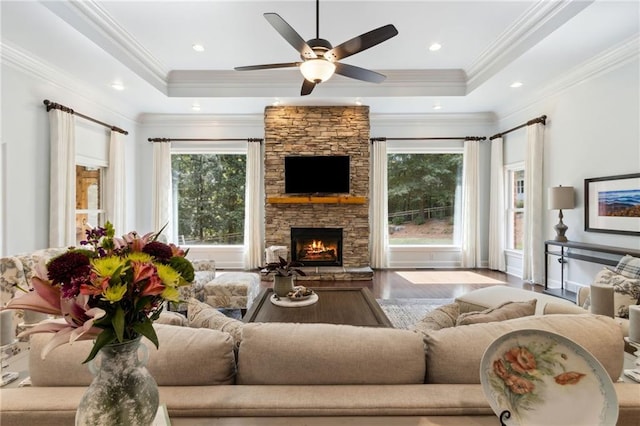 living room featuring a raised ceiling, a stone fireplace, wood-type flooring, crown molding, and ceiling fan