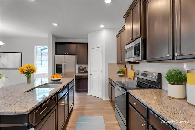 kitchen with light stone counters, stainless steel appliances, dark brown cabinets, light wood-type flooring, and a center island with sink