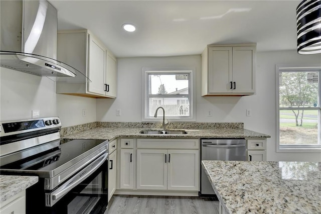 kitchen with a sink, wall chimney range hood, light wood-style flooring, light stone countertops, and appliances with stainless steel finishes