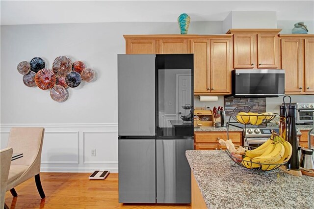 kitchen with wainscoting, light wood-style flooring, appliances with stainless steel finishes, light stone counters, and a decorative wall