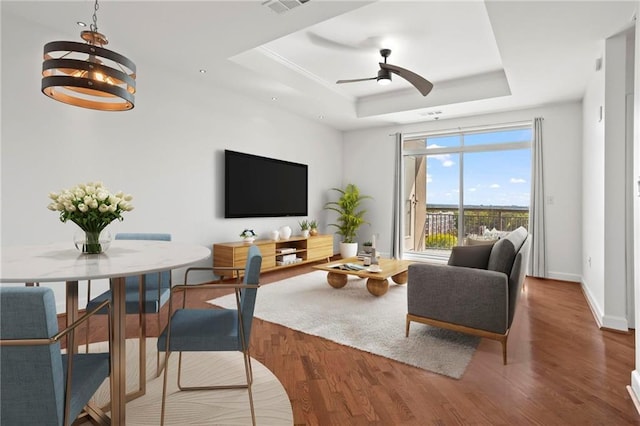 living area with ceiling fan with notable chandelier, a raised ceiling, baseboards, and wood finished floors