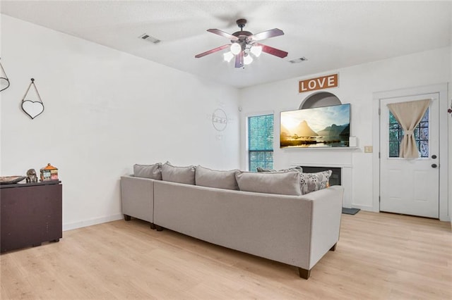 living area featuring light wood-style flooring, a fireplace, visible vents, baseboards, and a ceiling fan