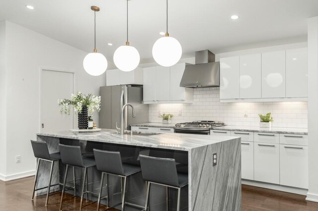 kitchen featuring a kitchen island with sink, white cabinetry, decorative light fixtures, and wall chimney exhaust hood