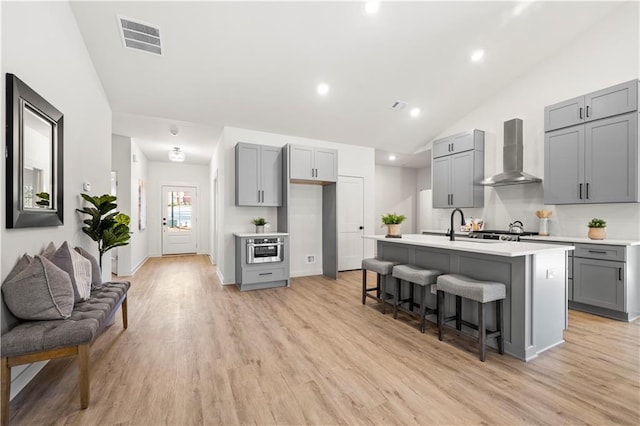 kitchen with a breakfast bar, a center island with sink, wall chimney range hood, gray cabinets, and light wood-type flooring