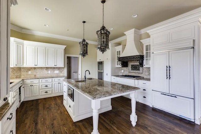 kitchen featuring custom exhaust hood, white cabinetry, built in appliances, decorative light fixtures, and a kitchen island with sink