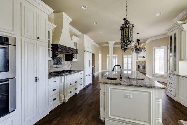 kitchen with ornate columns, appliances with stainless steel finishes, decorative light fixtures, an island with sink, and sink