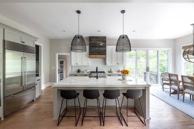 kitchen featuring sink, built in refrigerator, hanging light fixtures, light stone countertops, and a kitchen island with sink