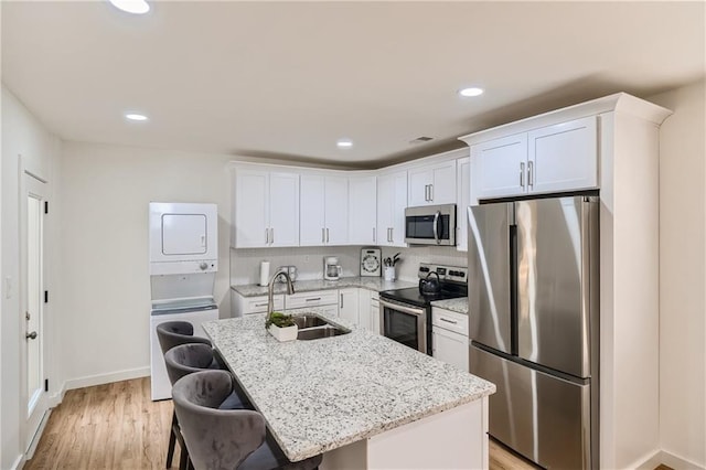 kitchen with stainless steel appliances, a sink, white cabinetry, stacked washer and clothes dryer, and a center island with sink