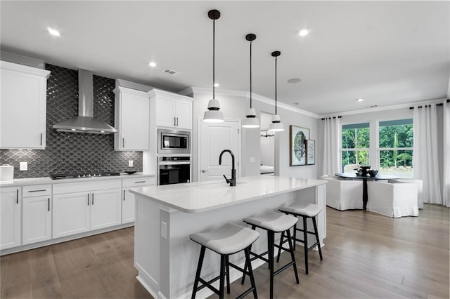 kitchen with stainless steel appliances, wall chimney range hood, white cabinets, and an island with sink