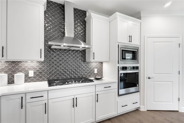 kitchen with wall chimney exhaust hood, backsplash, white cabinetry, light wood-type flooring, and appliances with stainless steel finishes