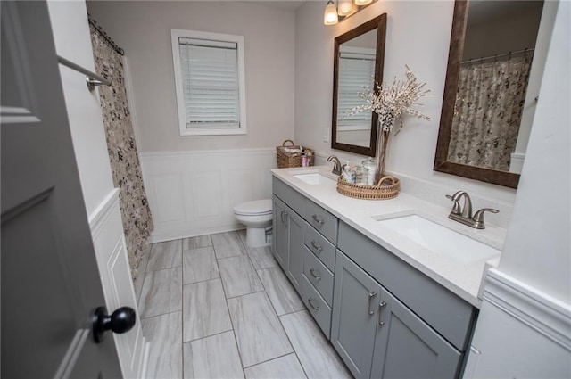 full bath with double vanity, toilet, a sink, and a wainscoted wall