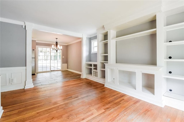 unfurnished living room featuring crown molding, a chandelier, built in shelves, wood finished floors, and a wainscoted wall