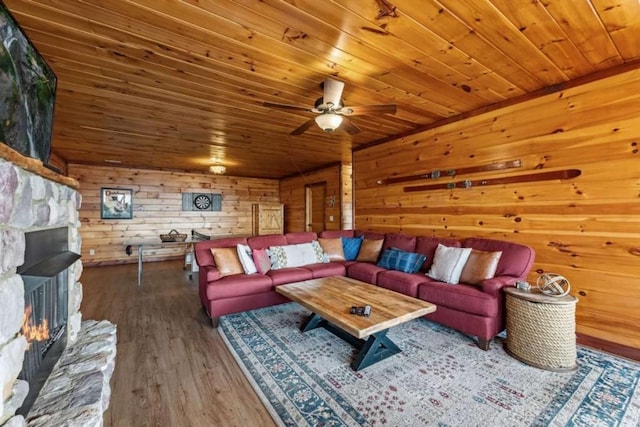 living room with wood finished floors, wooden walls, a stone fireplace, wooden ceiling, and ceiling fan