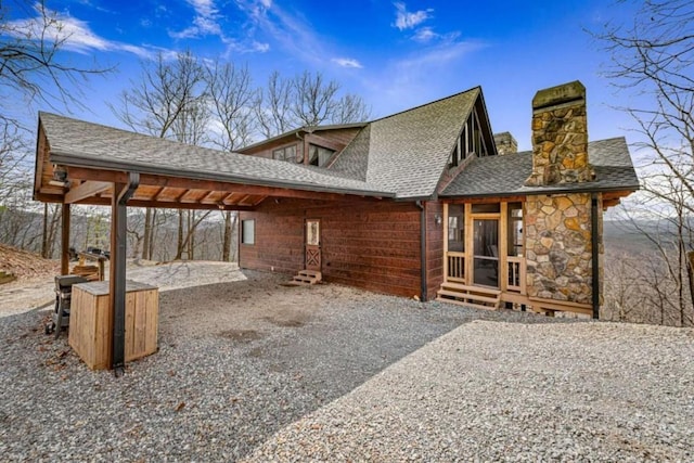 rear view of house featuring an attached carport, a shingled roof, entry steps, a chimney, and stone siding