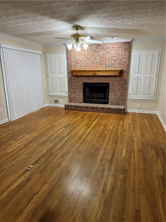 unfurnished living room featuring wood-type flooring, a textured ceiling, ceiling fan, crown molding, and a brick fireplace