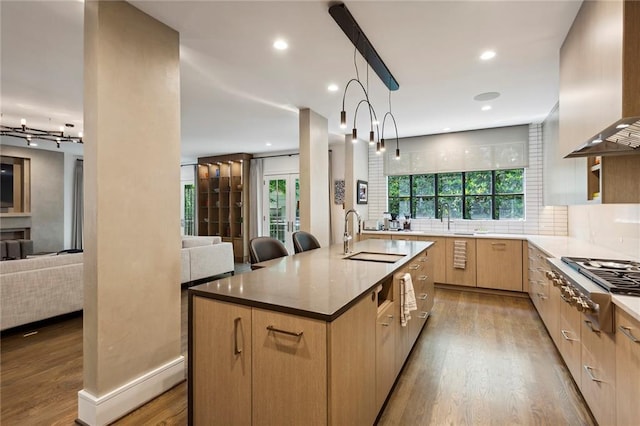 kitchen with open floor plan, wall chimney range hood, light brown cabinets, and stainless steel gas cooktop