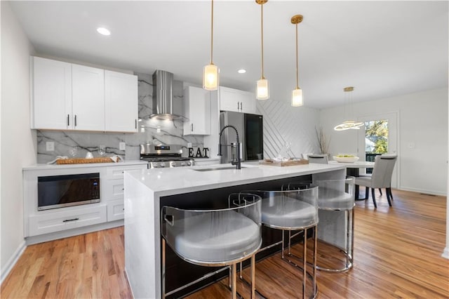 kitchen featuring a center island with sink, wall chimney exhaust hood, stainless steel appliances, and decorative light fixtures