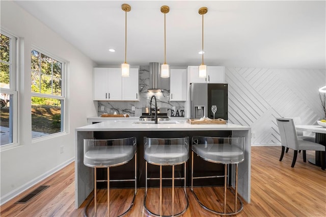 kitchen with white cabinetry, wall chimney exhaust hood, stainless steel fridge with ice dispenser, an island with sink, and pendant lighting