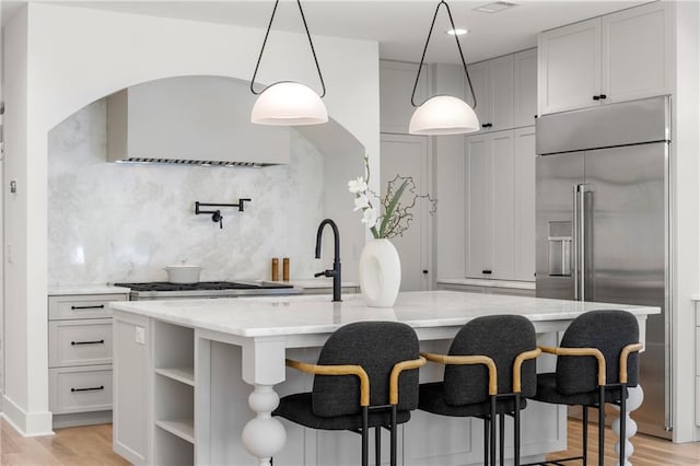 kitchen featuring an island with sink, built in refrigerator, pendant lighting, light wood-type flooring, and light stone counters