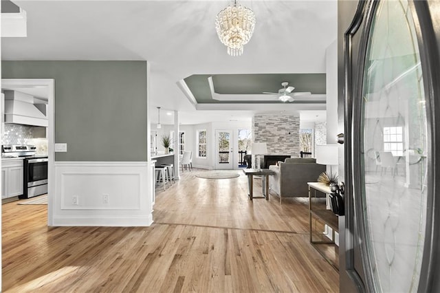 foyer with light wood-type flooring, a wainscoted wall, ceiling fan with notable chandelier, a stone fireplace, and a raised ceiling