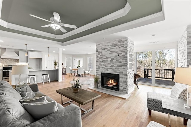 living area with a ceiling fan, light wood finished floors, a tray ceiling, a tiled fireplace, and crown molding
