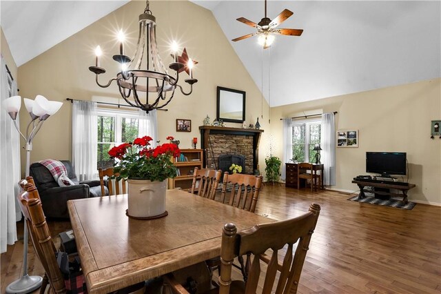 dining space with high vaulted ceiling, a stone fireplace, wood finished floors, and ceiling fan with notable chandelier