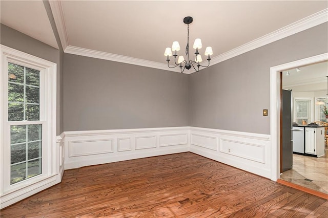 empty room with a chandelier, crown molding, wood finished floors, healthy amount of natural light, and wainscoting