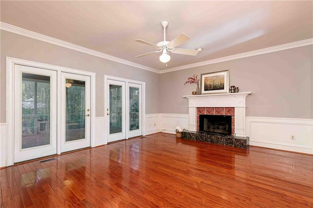 unfurnished living room with wood finished floors, a tiled fireplace, ornamental molding, a ceiling fan, and a wainscoted wall