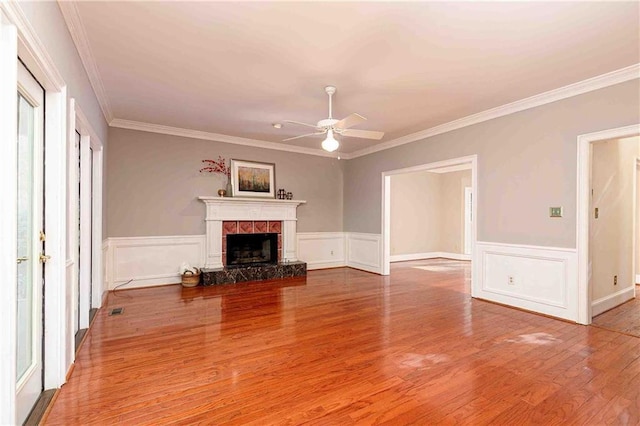 unfurnished living room with a ceiling fan, wood finished floors, ornamental molding, a fireplace, and a wainscoted wall