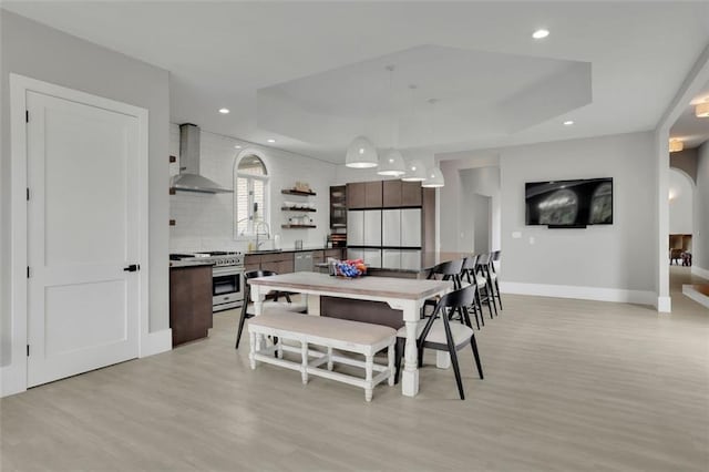 kitchen with stainless steel gas range oven, hanging light fixtures, wall chimney exhaust hood, dark brown cabinets, and light hardwood / wood-style floors