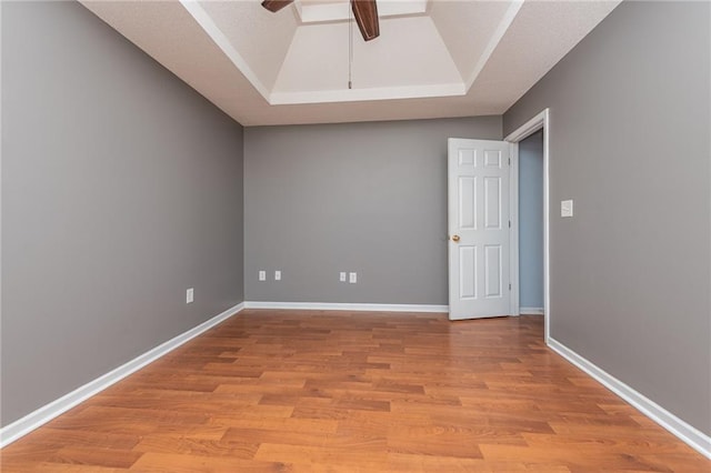 empty room with light wood finished floors, a ceiling fan, a raised ceiling, and baseboards