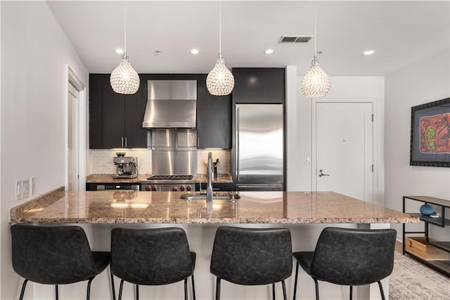 kitchen featuring built in refrigerator, hanging light fixtures, sink, and wall chimney range hood