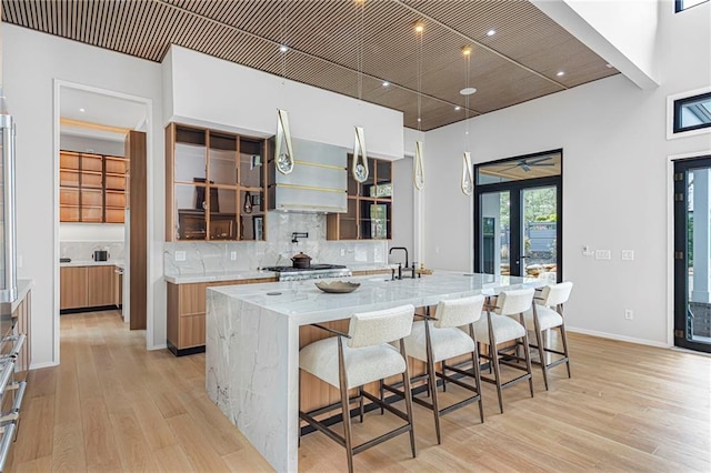 kitchen with wall chimney range hood, glass insert cabinets, backsplash, light wood-style floors, and a spacious island