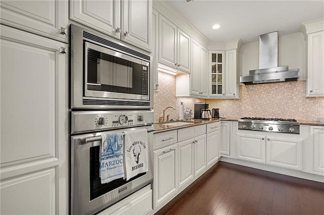 kitchen with dark wood-type flooring, wall chimney range hood, appliances with stainless steel finishes, stone countertops, and a sink