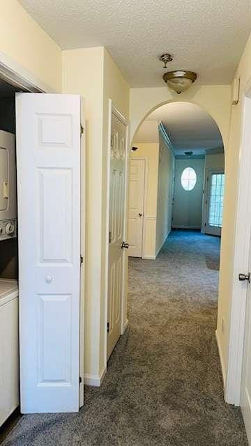 hall featuring stacked washer / drying machine, a textured ceiling, and dark colored carpet