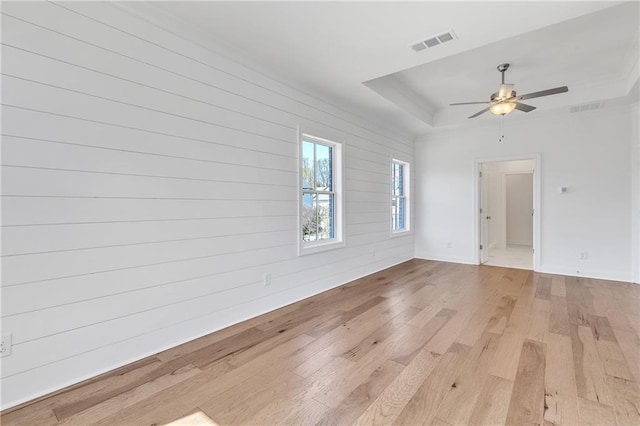 empty room with ceiling fan, wood walls, a tray ceiling, and light wood-type flooring