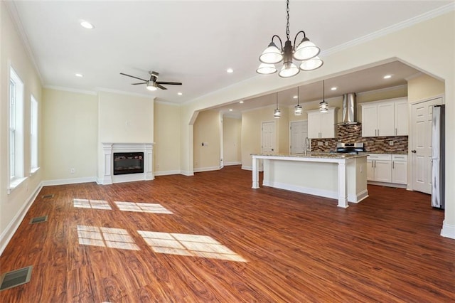 kitchen featuring decorative light fixtures, a center island with sink, white cabinetry, wall chimney exhaust hood, and ceiling fan with notable chandelier