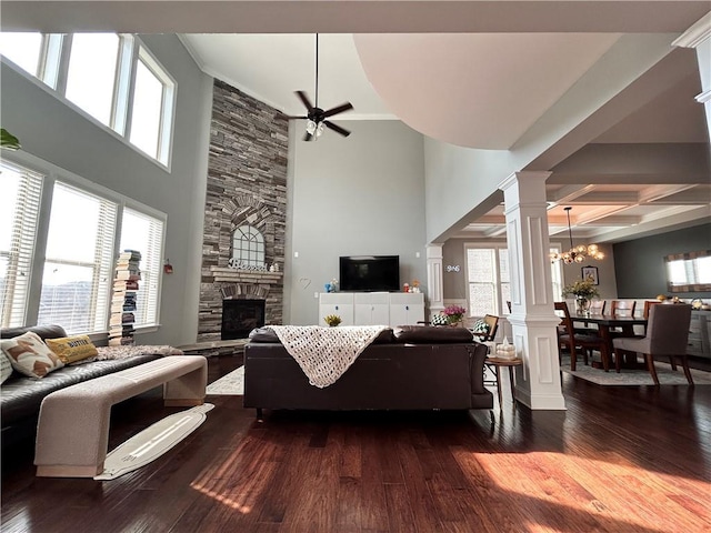 living room with a stone fireplace, ceiling fan with notable chandelier, a high ceiling, ornate columns, and dark wood-type flooring