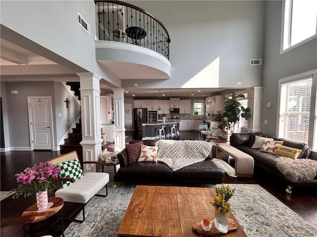 living area featuring ornate columns, dark wood-style flooring, a towering ceiling, and visible vents