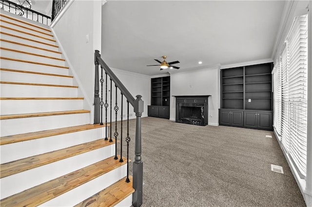 unfurnished living room with stairway, built in shelves, carpet floors, ceiling fan, and crown molding