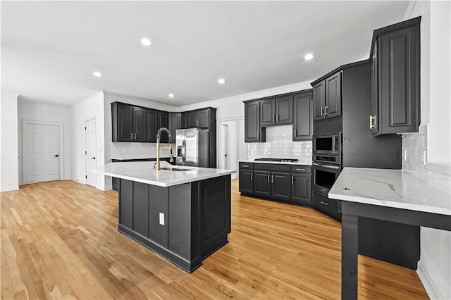 kitchen with a kitchen island with sink, light wood-style flooring, a sink, appliances with stainless steel finishes, and decorative backsplash