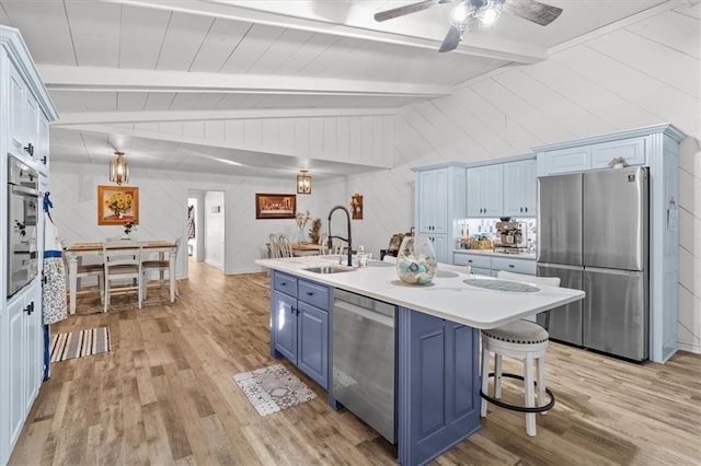 kitchen featuring vaulted ceiling with beams, blue cabinetry, an island with sink, appliances with stainless steel finishes, and light hardwood / wood-style floors