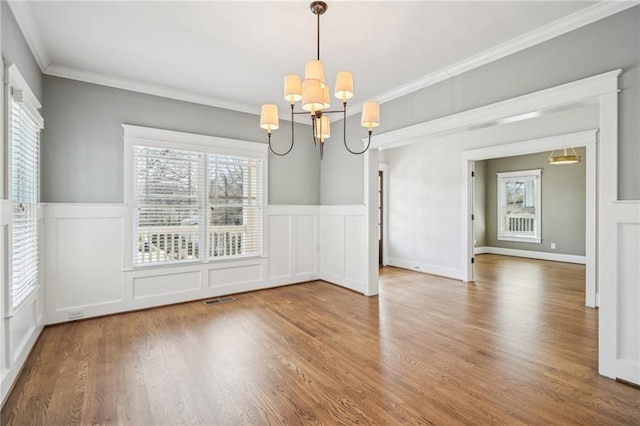 unfurnished dining area featuring wood finished floors, a wainscoted wall, visible vents, crown molding, and a chandelier