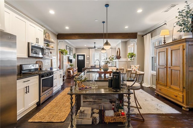 kitchen with hanging light fixtures, ornamental molding, appliances with stainless steel finishes, a kitchen island with sink, and white cabinets